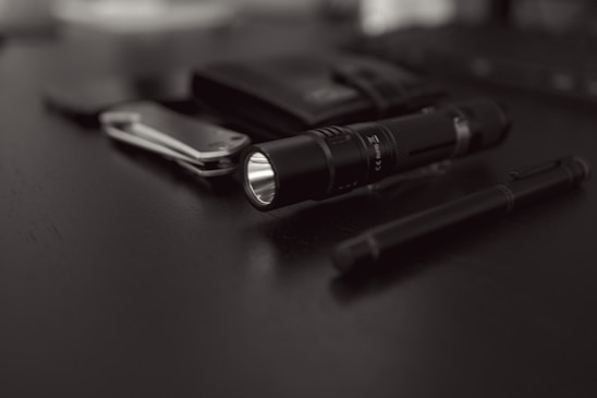 A collection of everyday carry items placed on a dark surface includes a flashlight, a folding knife, a pen, and a wallet. The focus is on the black flashlight with a textured grip, while the other items are slightly blurred in the background.