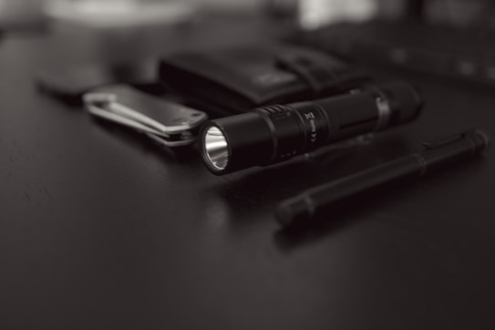 A collection of everyday carry items placed on a dark surface includes a flashlight, a folding knife, a pen, and a wallet. The focus is on the black flashlight with a textured grip, while the other items are slightly blurred in the background.