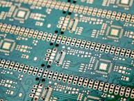 An intricate green circuit board with numerous white and black components, displaying a complex pattern of circuits, connections, and soldering points. The layout includes various microchips and connectors arranged in a repetitive and symmetrical design.