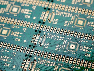 An intricate green circuit board with numerous white and black components, displaying a complex pattern of circuits, connections, and soldering points. The layout includes various microchips and connectors arranged in a repetitive and symmetrical design.