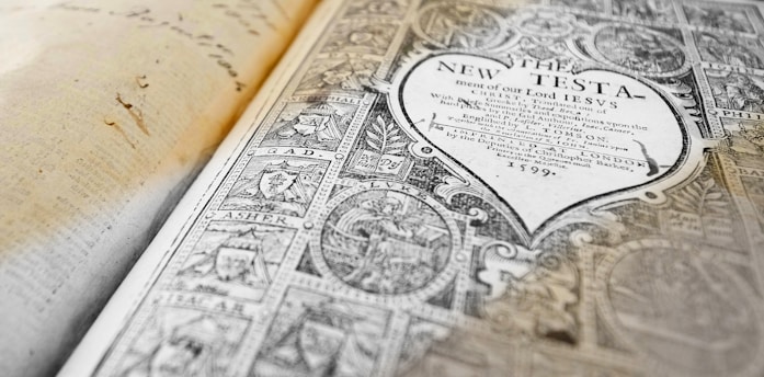 An open antique book displaying a detailed and ornate title page with intricate designs and text from 'The New Testament' dated 1599. The left side of the image shows an aged, handwritten page.
