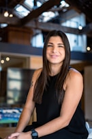 A woman with long dark hair is seated in a well-lit, modern interior with exposed beams and decorative lights. She is wearing a black sleeveless top and a watch, with a subtle smile on her face.