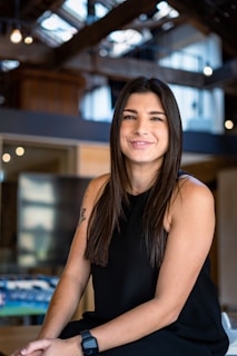 A woman with long dark hair is seated in a well-lit, modern interior with exposed beams and decorative lights. She is wearing a black sleeveless top and a watch, with a subtle smile on her face.
