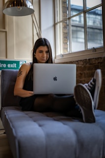 A woman with long dark hair is sitting comfortably on a couch, focused on her laptop. She is wearing a dark outfit and Converse sneakers. A floor lamp is positioned overhead, and there is a large window with a brick wall behind her. The setting suggests a modern, urban interior with exposed elements and natural light coming through the window.