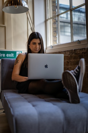 A woman with long dark hair is sitting comfortably on a couch, focused on her laptop. She is wearing a dark outfit and Converse sneakers. A floor lamp is positioned overhead, and there is a large window with a brick wall behind her. The setting suggests a modern, urban interior with exposed elements and natural light coming through the window.