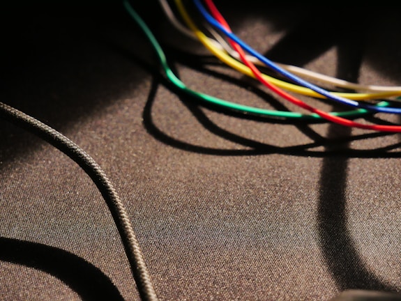 Various colorful cables, including blue, green, yellow, red, and white, are intertwined and cast shadows on a textured dark surface. A thicker black cable moves diagonally across the lower left corner.