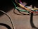 Various colorful cables, including blue, green, yellow, red, and white, are intertwined and cast shadows on a textured dark surface. A thicker black cable moves diagonally across the lower left corner.