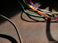 Various colorful cables, including blue, green, yellow, red, and white, are intertwined and cast shadows on a textured dark surface. A thicker black cable moves diagonally across the lower left corner.