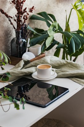 A cozy scene featuring a white ceramic cup filled with coffee placed on a saucer on a white table. Nearby, an electronic tablet with a black screen sits alongside a green cloth. A black vase holds dried brown foliage and green leafy plants add a touch of nature to the setup. The background is a textured beige wall, creating a warm and inviting atmosphere.