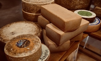 A collection of variously shaped cheeses is placed on a wooden surface, with circular and rectangular blocks prominently featured. The cheeses display a rough and aged texture, with some labels partially visible.