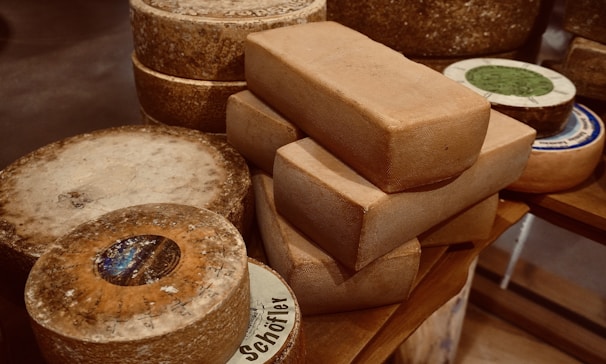 A collection of variously shaped cheeses is placed on a wooden surface, with circular and rectangular blocks prominently featured. The cheeses display a rough and aged texture, with some labels partially visible.
