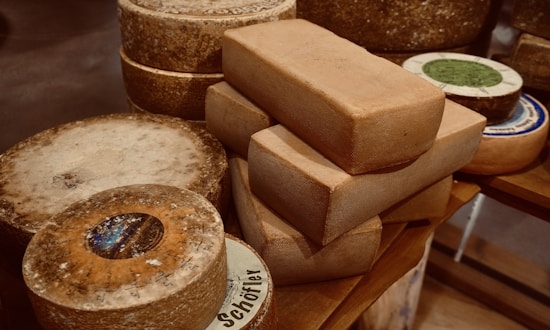 A collection of variously shaped cheeses is placed on a wooden surface, with circular and rectangular blocks prominently featured. The cheeses display a rough and aged texture, with some labels partially visible.