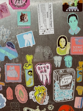 A collection of colorful stickers on a textured surface, featuring various designs including a cat photo with text, a tooth with 'BITE ME' text, artistic portraits, a packet labeled 'Ms. Toof Ache&rsquo;s Toof Tac,' and several abstract and cartoon-like images.