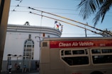 A trolleybus labeled 'Clean Air Vehicle' is passing by a building with large arched windows. Several people are waiting at a bus stop under an awning. Colorful flags adorn the top of the building, and palm tree leaves extend into the scene from the right. Overhead wires and urban infrastructure are visible.