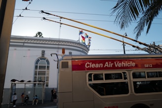 A trolleybus labeled 'Clean Air Vehicle' is passing by a building with large arched windows. Several people are waiting at a bus stop under an awning. Colorful flags adorn the top of the building, and palm tree leaves extend into the scene from the right. Overhead wires and urban infrastructure are visible.