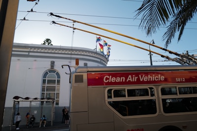 A trolleybus labeled 'Clean Air Vehicle' is passing by a building with large arched windows. Several people are waiting at a bus stop under an awning. Colorful flags adorn the top of the building, and palm tree leaves extend into the scene from the right. Overhead wires and urban infrastructure are visible.