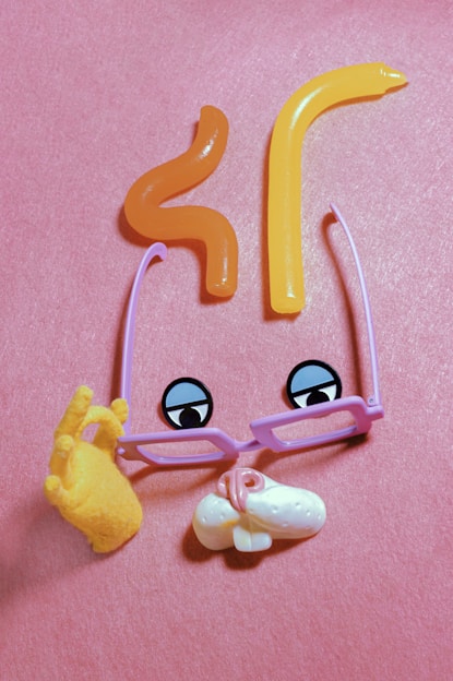 A whimsical and playful composition using various objects arranged to resemble a face. There are two jelly-like orange shapes forming the eyebrows, purple eyeglass frames with cartoonish eyes, a yellow hand making an OK gesture, and a white object representing a nose with a small pink detail suggesting a tongue. The pink background adds a vibrant and cheerful setting.
