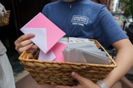 A person holds a wicker basket filled with white and pink envelopes, wearing a blue t-shirt with a logo of a church in New York.