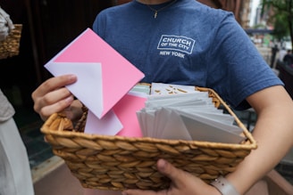 A person holds a wicker basket filled with white and pink envelopes, wearing a blue t-shirt with a logo of a church in New York.