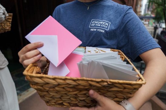 A person holds a wicker basket filled with white and pink envelopes, wearing a blue t-shirt with a logo of a church in New York.
