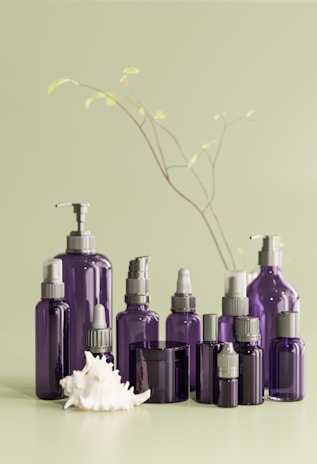 A collection of purple cosmetic bottles of various shapes and sizes are arranged neatly on a light green surface. The bottles feature a mix of caps and pump dispensers. In the foreground, a large white seashell is prominently displayed. Delicate branches with small leaves extend in the background, adding an organic touch.