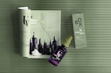 Green and violet-themed display includes a magazine spread showcasing cosmetic bottles, with a spray bottle and its packaging box arranged on top. The textured background and the minimalist design elements highlight the violet glass bottles and the elegant layout.