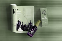Green and violet-themed display includes a magazine spread showcasing cosmetic bottles, with a spray bottle and its packaging box arranged on top. The textured background and the minimalist design elements highlight the violet glass bottles and the elegant layout.