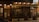 A quaint cafe facade featuring wooden paneling and vintage signs that read 'Cafe Riche.' Warm glowing lights illuminate the exterior, and potted plants add a touch of greenery. The entrance is adorned with glass doors and windows showcasing various posters and notices.