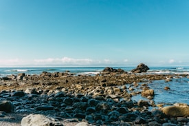 A serene coastal landscape featuring a rocky shoreline with a clear blue sky overhead. Ocean waves gently crash against the rocks in the foreground. The horizon showcases a smooth transition between the ocean and sky, with distant clouds adding depth to the scene.