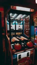 An indoor basketball arcade game with a retro design, featuring two hoops, multiple basketballs, and score displays. The game is illuminated by overhead lights, casting a vintage ambiance.
