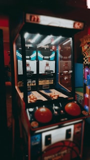An indoor basketball arcade game with a retro design, featuring two hoops, multiple basketballs, and score displays. The game is illuminated by overhead lights, casting a vintage ambiance.