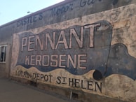 A weathered outdoor mural painted on the side of a building advertises 'Pennant Kerosene' with additional text 'Local Depot St Helen.' The mural features large, bold lettering and uses a scroll-like design. The wall shows signs of age, with faded paint and a rustic appearance, contributing to a vintage feel.