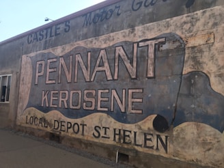 A weathered outdoor mural painted on the side of a building advertises 'Pennant Kerosene' with additional text 'Local Depot St Helen.' The mural features large, bold lettering and uses a scroll-like design. The wall shows signs of age, with faded paint and a rustic appearance, contributing to a vintage feel.
