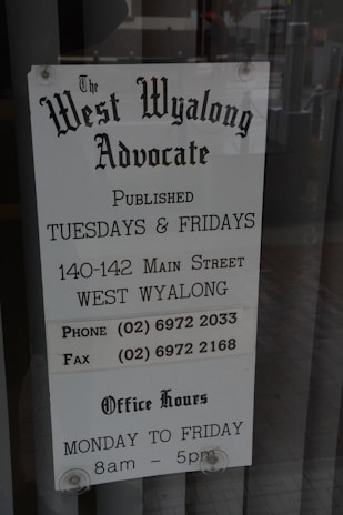 A sign for 'The West Wyalong Advocate' newspaper, detailing publication days as Tuesdays and Fridays. The sign includes the street address, telephone and fax numbers, and office hours from Monday to Friday, 8 am to 5 pm. The background consists of a reflective glass surface with metal rivets at the top corners.