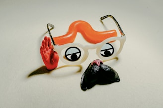 A playful composition featuring novelty glasses with white frames, cartoon-style eyes, and an attached orange curly toy mustache. A red hand is positioned in a waving gesture beside the glasses.