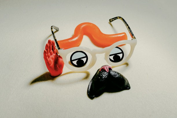 A playful composition featuring novelty glasses with white frames, cartoon-style eyes, and an attached orange curly toy mustache. A red hand is positioned in a waving gesture beside the glasses.