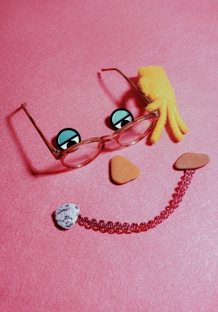 A playful arrangement of objects on a pink surface, including a pair of glasses with cartoonish eyes drawn on the lenses, an orange plush crab toy, and various stones and beads forming a smiling face.