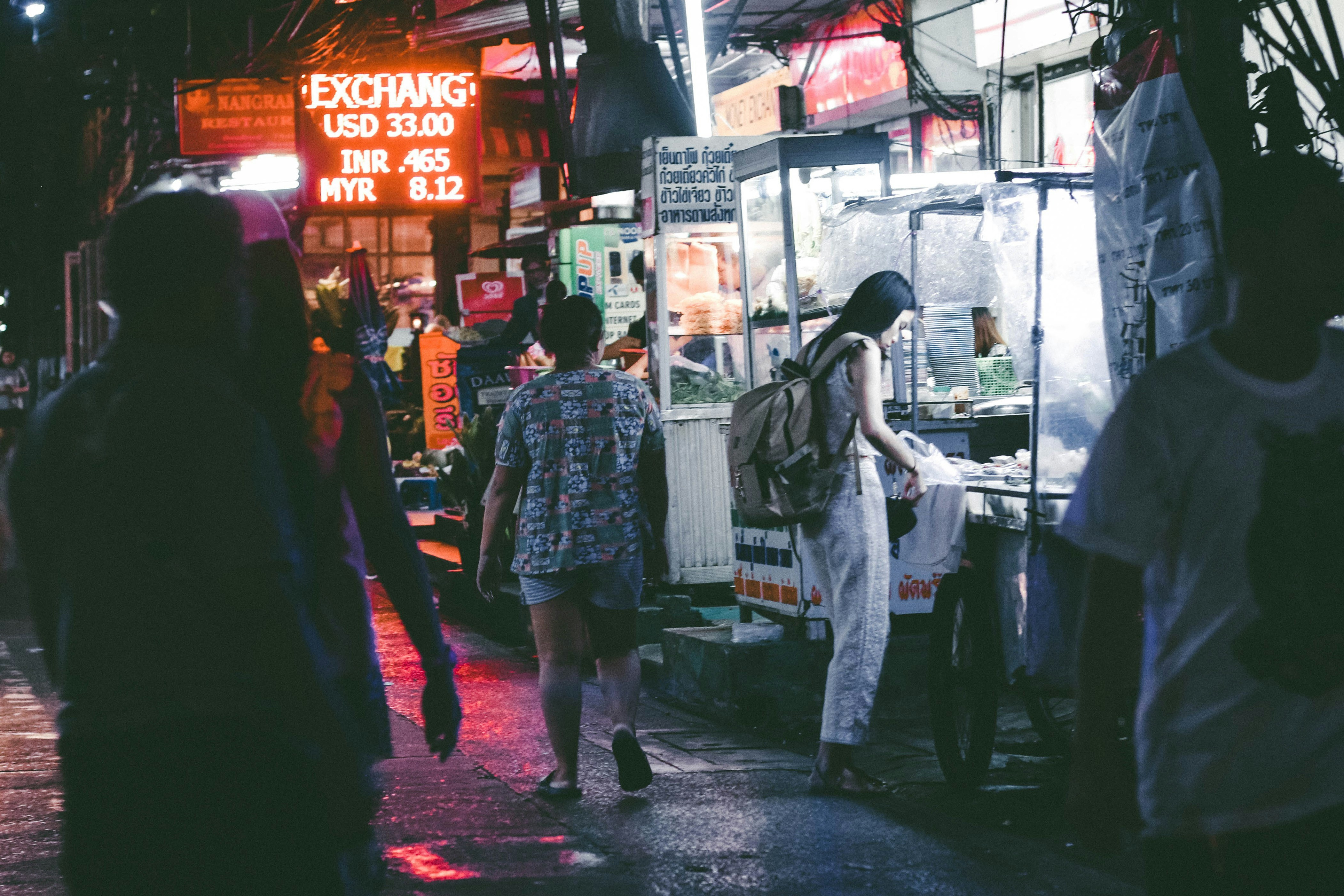 a group of people walking down a street at night, A woman is standing in front of a street food stall