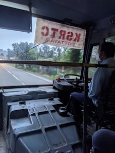 A bus interior with the driver sitting at the front right corner, visible through a window shield. The road ahead is visible through a large windshield adorned with a red and white sign. Trees and roadside scenery are visible outside.