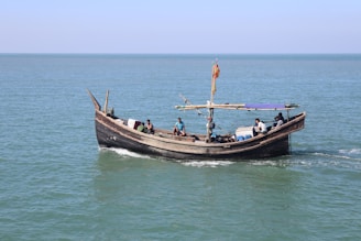 A small wooden boat with an arched hull sails on a calm sea. It is manned by several individuals engaged in various activities aboard the vessel, with one person navigating. A colorful flag is attached to a pole in the center, and several items or containers are visible on the deck.