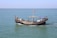 A small wooden boat with an arched hull sails on a calm sea. It is manned by several individuals engaged in various activities aboard the vessel, with one person navigating. A colorful flag is attached to a pole in the center, and several items or containers are visible on the deck.