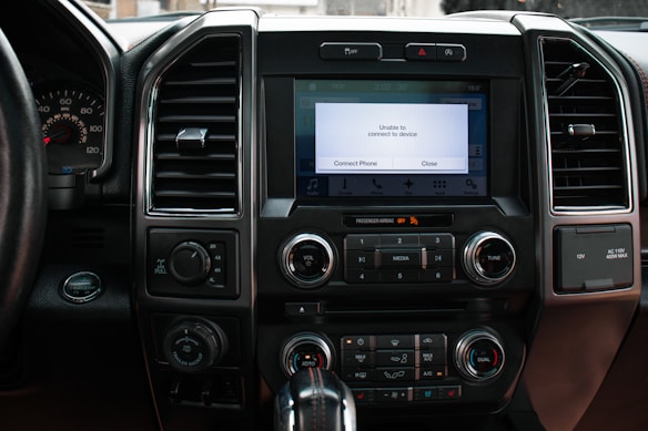 The interior of a vehicle dashboard featuring a central infotainment screen displaying a message indicating the inability to connect to a device. Surrounding the screen are various buttons and controls for air conditioning, media, and vehicle settings. The dashboard includes air vents, a steering wheel to the left, and several knobs and dials for adjusting settings.
