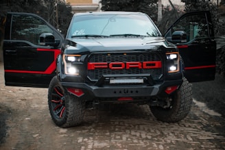 A rugged black pickup truck with bold red accents on the grill and rims is parked outdoors on a dirt road. The truck has both front doors open, revealing the darkness inside. The background includes greenery and a building partially visible in the distance, providing a sense of an outdoor or rural setting.