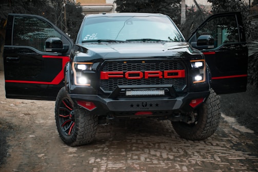 A rugged black pickup truck with bold red accents on the grill and rims is parked outdoors on a dirt road. The truck has both front doors open, revealing the darkness inside. The background includes greenery and a building partially visible in the distance, providing a sense of an outdoor or rural setting.
