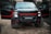 A rugged black pickup truck with bold red accents on the grill and rims is parked outdoors on a dirt road. The truck has both front doors open, revealing the darkness inside. The background includes greenery and a building partially visible in the distance, providing a sense of an outdoor or rural setting.
