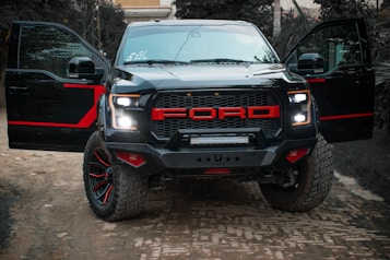 A rugged black pickup truck with bold red accents on the grill and rims is parked outdoors on a dirt road. The truck has both front doors open, revealing the darkness inside. The background includes greenery and a building partially visible in the distance, providing a sense of an outdoor or rural setting.
