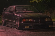 A dark, sleek car, possibly a BMW, is parked on a wet surface. The image is taken at night, and the car is illuminated by soft lighting, highlighting its glossy exterior. The setting appears to be outdoors with trees faintly visible in the background. A custom windshield banner is visible.