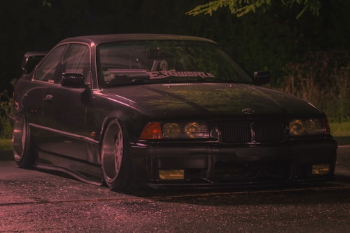 A dark, sleek car, possibly a BMW, is parked on a wet surface. The image is taken at night, and the car is illuminated by soft lighting, highlighting its glossy exterior. The setting appears to be outdoors with trees faintly visible in the background. A custom windshield banner is visible.