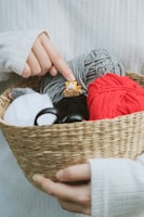 A woven basket held in two hands contains several balls of yarn in various colors, including red, gray, white, and black. On top of the yarn, a small figurine of a yellow and white animal with small ears and eyes sits, partially in a small cardboard box. The index finger of the right hand is pointing towards this figurine. The person holding the basket is wearing a white ribbed sweater.