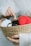 A woven basket held in two hands contains several balls of yarn in various colors, including red, gray, white, and black. On top of the yarn, a small figurine of a yellow and white animal with small ears and eyes sits, partially in a small cardboard box. The index finger of the right hand is pointing towards this figurine. The person holding the basket is wearing a white ribbed sweater.